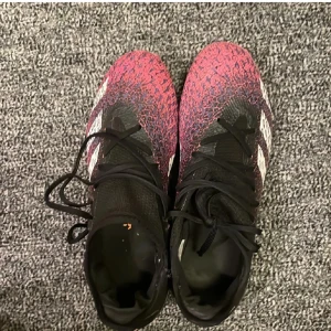 Adidas Predator fotbollsskor svart/röd - Säljer ett par Adidas Predator fotbollsskor med coolt mönster i svart, rött och lila. Skorna har snörning, tre vita ränder på sidorna och en texturad ovandel för extra grepp. Sulan är röd och lila med dobbar för gräsplan. Perfekta för dig som vill sticka ut på planen!