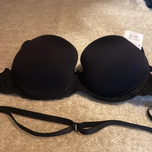 Svart push up bh  från  Kris Line 80DD - Svart push up  Bh från Kris Line med släta kupor, avtagbara axelband och spetsdetaljer längs nederkanten. Söt liten rosett framtill. Perfekt under axelbandslösa plagg och med justerbara band för bästa passform.