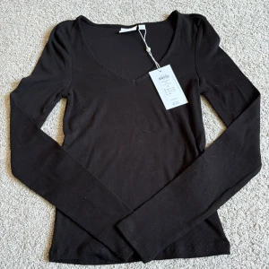 Svart ribbad långärmad topp från Vila - Svart långärmad topp från Vila i ribbad bomull med snygg v-ringning. Modellen är slim och har en enkel, stilren look som funkar till många olika outfits. Perfekt för dig som gillar basic med en twist. Storlek XS.