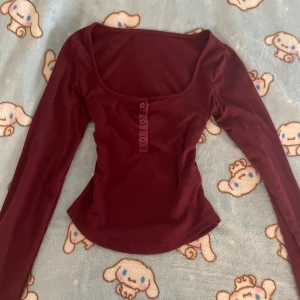 Vinröd urringad Elena Gilbert långärmad topp  - Snygg vinröd långärmad topp med ribbad struktur och bred, rundad urringning. Framtill finns tre knappar som detalj. Modellen har en mjuk, stretchig känsla. Perfekt för dig som gillar stilrena och enkla plagg med en twist. Storlek M men tröjan är mer som S-XS.