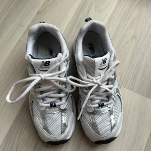 Vita New Balance 530 sneakers strl 38 - Säljer ett par klassiska vita New Balance sneakers i storlek 38. Skorna har meshpaneler och detaljer i silver, med snörning och rund tå. Perfekta för dig som gillar sportig och clean stil. Ikonisk N-logga på sidan och bekväm sula för vardagsbruk.