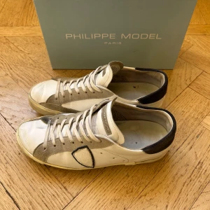 Philippe Model vita sneakers - Snygga sneakers från Philippe Model i vitt och grått med svarta detaljer. Skorna har en rund tå, platt sula och snörning. Ovandelen är i skinn och mocka, med en kontrasterande svart häl och en dekorativ patch på sidan. Perfekt för dig som gillar stilrena och trendiga sneakers.