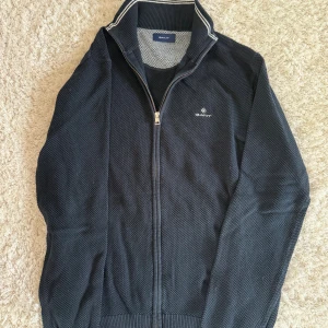 Mörkblå kofta från GANT med dragkedja - Säljer en stilren mörkblå kofta från GANT i storlek L. Koftan har hel dragkedja, ribbade muddar och krage med vita ränder. Materialet är stickat och ger en snygg struktur. Perfekt för lager-på-lager och passar till många outfits.