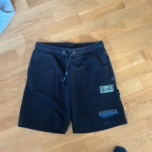 Svarta shorts från Hoodrich med tryck ( storlek L i barn) har även XS i vuxen!!! - Svarta shorts från Hoodrich med blå detaljer och tryck på framsidan. De har snörning i midjan och två sidofickor. Materialet är mjukt och skönt, perfekt för chill eller streetwear. Snygga loggor och patch framtill ger en modern vibe.