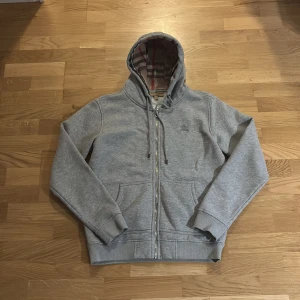 Grå hoodie från Burberry med dragkedja - Grå hoodie från Burberry med klassiskt rutigt foder i huvan. Tröjan har dragkedja framtill, snörning i huvan och två fickor. Tillverkad i mjuk bomull med en liten broderad logga på bröstet. Perfekt för en chill och stilren look.