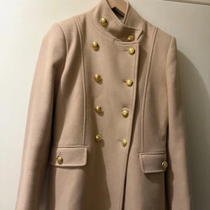 Beige kappa med guldknappar från Zara - Stilren beige kappa från Zara med snygga guldknappar i dubbelknäppt design och hög krage. Jackan har markerade axlar, ficklock och är tillverkad i ullblandning från Manteco. Perfekt för dig som vill ha en klassisk och trendig look.