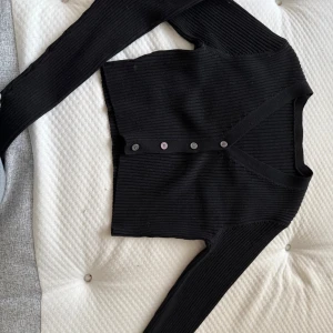 Svart ribbad kofta med knappar - Svart ribbad kofta med v-ringning och fyra knappar framtill. Långärmad och croppad modell som sitter snyggt på kroppen🩷