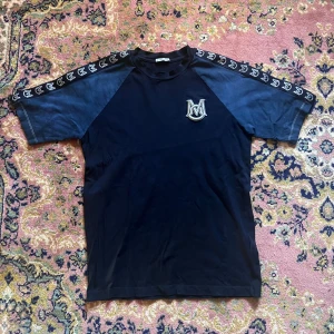 Svart t-shirt med logga från Moncler M - Snygg svart t-shirt från Moncler med kontrasterande mörkblå ärmar och coola loggor längs axlarna. På bröstet sitter en broderad Moncler-logga i vitt. Perfekt för dig som gillar streetwear och vill sticka ut lite extra.