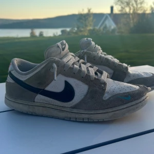 Nike Dunk Low i vitt och grått. Sänkbart pris - Nike Dunk Low sneakers i vitt och grått med blå swoosh och röd Nike-logga på hälen. Ovandel i skinn och mocka, klassisk snörning och platt sula. Perfekta för dig som gillar retrovibbar och streetstyle.