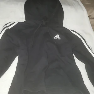 Svart Adidas hoodie  - Svart hoodie från Adidas med klassiska vita ränder längs ärmarna och Adidas-logga på bröstet. Tröjan har huva med snörning och är långärmad. Perfekt för en sportig och avslappnad stil.