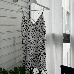 Vit leopardmönstrad klänning H&M - Säljer en vit kortklänning från H&M med svart leopardmönster. Klänningen har tunna axelband som korsas i ryggen och en rak, avslappnad passform. 