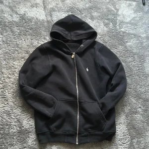 Svart hoodie från Polo Ralph Lauren - Svart hoodie från Polo Ralph Lauren med dragkedja och huva. Klassisk design med liten vit logga på bröstet, två fickor fram och ribbade muddar. Perfekt för chill dagar och enkel att matcha med jeans eller joggers.