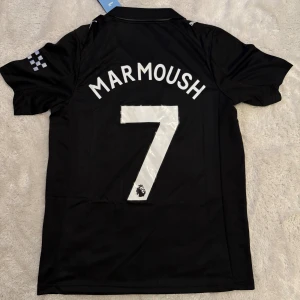 Svart Manchester City Marmoush tröja - Snygg svart Manchester City fotbollströja från Puma med Marmoush och nummer 7 på ryggen. Tröjan har korta ärmar, klassisk krage och silverfärgade detaljer som klubbmärke och Etihad Airways-logga. Tillverkad i lätt och ventilerande material för maximal komfort.