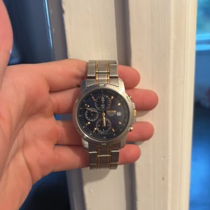Seiko  herrklocka  - Säljer en snygg Seiko kronograf med rund boett och länkarmband i silverfärgat stål med guldfärgade detaljer. Urtavlan är mörkblå med tre subdials och datumvisning. Klockan har tydliga knappar i guld på sidan och klassisk Seiko-gravyr på baksidan.