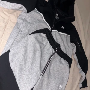 Nike Tech Fleece  - Nike hoodie i svart och ljusgrå med vit detalj över bröstet. Tröjan har huva, lång ärm och dragkedja framtill. Materialet är mjuk bomull och polyester, perfekt för chill eller träning. Snyggt Nike-logo på bröstet och cool design med kontrastpaneler.