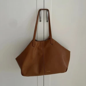 Brun skinnväska från Polène - Säljer en snygg och rymlig brun axelväska från Polène i mjukt skinn. Väskan har stilren design med två långa handtag och diskret logga framtill. Perfekt för dig som vill ha plats för allt du behöver under dagen. Använt skick men har mycket kvar att ge. 