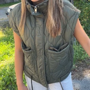 Zara väst - Snygg olivgrön dunväst med quiltad design, hög krage och dragkedja framtill. Västen har två stora fickor och dragsko i nederkant för extra passform. Perfekt att slänga över en hoodie eller tröja för en cool lager-på-lager-look.