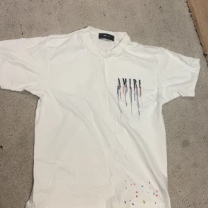 Vit AMIRI t-shirt med färgstänk - Säljer en vit t-shirt från AMIRI med coolt tryck och färgstänk både fram och bak. Stora AMIRI-bokstäver med droppande färgdetaljer ger en unik vibe. Klassisk passform och rund hals. Perfekt för dig som vill sticka ut med streetstyle.