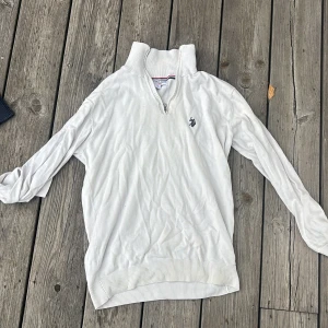Vit half zip-tröja från Polo Assn - Vit långärmad tröja från U.S. Polo Assn med half zip och hög krage. Tröjan har broderad logga på bröstet och ribbade muddar vid ärmslut och nederkant. Perfekt för en clean och sportig stil.