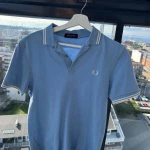 Blå pikétröja från Fred Perry - Snygg ljusblå pikétröja från Fred Perry med klassisk krage och vita ränder på krage och ärmslut. Tröjan har korta ärmar och ikonisk Fred Perry-logga broderad på bröstet. Perfekt för en stilren och avslappnad look.