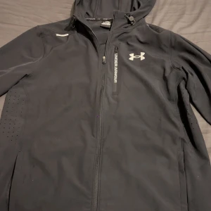 Svart vindjacka från Under Armour - Svart vindjacka från Under Armour med huva och dragkedja framtill. Jackan har en bröstficka med dragkedja och logga på bröstet. Lätt material och diskreta ventilationshål på sidorna gör den perfekt för aktiva dagar.