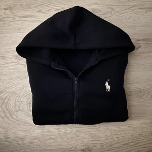 Ralph lauren zip - Svart hoodie från Polo Ralph Lauren med klassisk vit logga på bröstet. Tröjan har huva, dragkedja framtill och långa ärmar. Tillverkad i mjuk bomull, perfekt för chill dagar. Snygg och enkel design som funkar året runt.