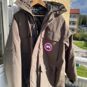 Canada Goose jacka - Säljer en brun Canada Goose dunjacka med stor huva och avtagbar pälskant.  Nypris 17000 kr. Jackan är äkta