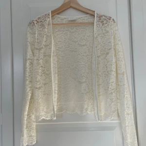 Vit spetsig kofta från Vila - Säljer en superfin vit kofta från Vila i genombruten spets med blommigt mönster. Toppen har lång ärm, öppen framtill och är perfekt att slänga över ett linne eller klänning för en extra touch. Materialet är mjukt och stretchigt syntet.