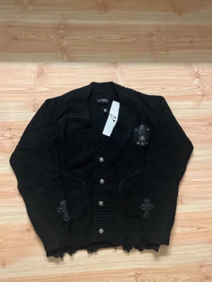 Chrome Hearts button up - Säljer en svart chunky stickad kofta från Chrome Hearts med grova ribbade muddar, dekorativa silverknappar och läderpatches med korsdetaljer. Koftan har två fickor framtill och råa kanter nertill för en edgy vibe. Perfekt för dig som gillar statement-plagg.