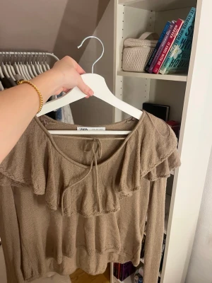 Beige volangtröja från Zara - Supersöt beige tröja från Zara med volangdetaljer över axlar och bröst samt knyt i halsen. Långärmad och i ett tunt stickat material som känns luftigt men ändå mysigt. Perfekt för dig som gillar romantiska vibes och vill ha något extra till din outfit.