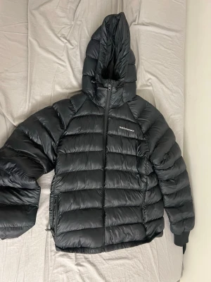 Svart pufferjacka Peak Performance L - Säljer en svart pufferjacka från Peak Performance i storlek L. Jackan har huva, dragkedja framtill och två sidofickor. Den är quiltad och har en clean look med diskret logga på bröstet. Perfekt för kalla dagar och riktigt skön att bära. Helt ny köpte present till min mamma men den passade inte henne 