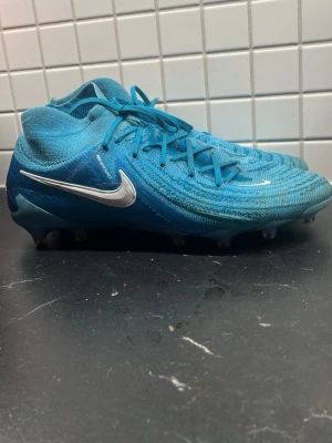 Nike Phantom Luna Elite 43 AG - Säljer ett par blå Nike fotbollsskor med vit swoosh och snörning. Skorna har stickad ovandel och högre strumpliknande skaft för extra stöd. Sulan är utrustad med fasta dobbar för konstgräsplan. Perfekta för dig som vill sticka ut på planen. Varken påse eller orginalbox tillkommer ⚠️