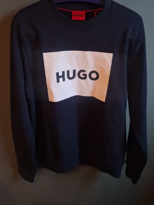 Svart sweatshirt från HUGO med logga - Säljer en svart sweatshirt från HUGO med stor vit logga framtill. Tröjan har rund halsringning och långa ärmar. Tillverkad i mjuk bomull för skön känsla. Perfekt för dig som gillar stilrena och trendiga plagg, stl XS så tog storleken 34. 