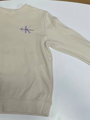 Beige sweatshirt från Calvin Klein Jeans - Snygg beige sweatshirt från Calvin Klein Jeans med lila CK-logga och svart text på bröstet. Tröjan har rund hals, ribbade muddar och är långärmad. Perfekt för en clean och stilren look.