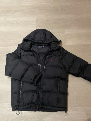 Mörkblå pufferjacka från Polo Ralph Lauren - Säljer en murkblå pufferjacka från Polo Ralph Lauren med huva och dragkedja. Jackan har två sidofickor med dragkedja och den klassiska röda Polo-loggan broderad på bröstet. Perfekt för kalla dagar och riktigt snygg till vintern! 🤩 Man kan även spenna åt midjan på jackan. 