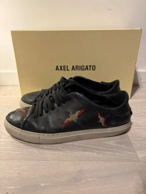Svarta Axel Arigato sneakers  - Snygga svarta sneakers från Axel Arigato i skinn med broderade fåglar i rött, blått och beige på sidan och tån. Klassisk låg modell med vita gummisulor och svarta snören. Perfekta för dig som vill ha en unik touch på dina skor.