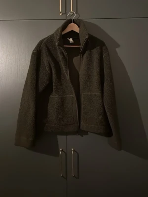 Mörkbrun teddyjacka med dragkedja - Mysig mörkgrön oversized teddyjacka med hög krage och två stora fickor framtill från H&M. Jackan har en rak passform och stängs med dragkedja. Perfekt för kyliga dagar när du vill vara både varm och stilren. 