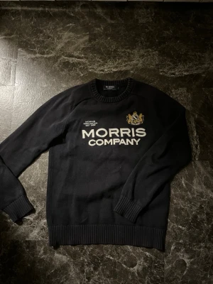 Svart stickad tröja från Morris - Säljer en svart stickad tröja från Morris Company med broderad logga och text framtill. Tröjan har rund halsringning, ribbade muddar och en klassisk passform. Perfekt för dig som gillar stilrena och tidlösa plagg.