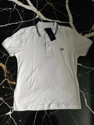 Vit piké från Fred Perry - Klassisk vit pikétröja från Fred Perry med svarta ränder på krage och ärmslut. Tröjan har korta ärmar, knappar framtill och den ikoniska Fred Perry-loggan broderad på bröstet. Tillverkad i mjuk bomull som andas.