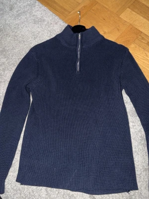Mörkblå stickad half zip-tröja 157 - Mörkblå ribbstickad tröja från 157 med half zip-dragkedja i halsen. Tröjan har hög krage och långa ärmar, perfekt för kyligare dagar. Klassisk och enkel design som funkar till det mesta.