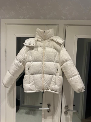 Vit dunjacka från Moncler - Säljer en snygg vit pufferjacka från Moncler med hög krage och avtagbar huva. Jackan har dragkedja framtill, två fickor med dragkedja och klassisk Moncler-logga på ärmen. Perfekt för kalla dagar och riktigt trendig look.