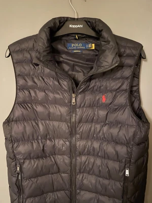 Svart dunväst från Polo Ralph Lauren - Säljer en svart dunväst från Polo Ralph Lauren med quiltad design och hög krage. Västen har dragkedja framtill, två sidofickor med dragkedja och den klassiska röda loggan broderad på bröstet. Perfekt för lager-på-lager och riktigt snygg till höst och vinter. Perfekt skick