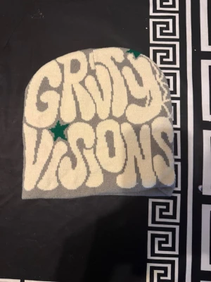 Groovy Visions mössa i olika färger - Säljer stickade mössor med texten 'Groovy Visions' i retrostil. Finns i grå/vit med gröna stjärnor, blå/orange, lila/gul och röd/vit. Mössorna har coolt mönster och är tillverkade i mjukt syntetmaterial. Perfekt accessoar för att sticka ut i mängden.