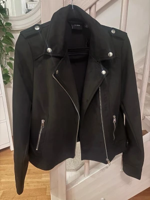 Svart bikerjacka från VERO MODA - Snygg svart bikerjacka från VERO MODA med silvriga dragkedjor och knappar. Jackan har klassisk krage, axelklaffar och två fickor med dragkedja. Materialet är mjukt och matt, vilket ger en stilren look. Perfekt för dig som vill ha en edgy vibe.