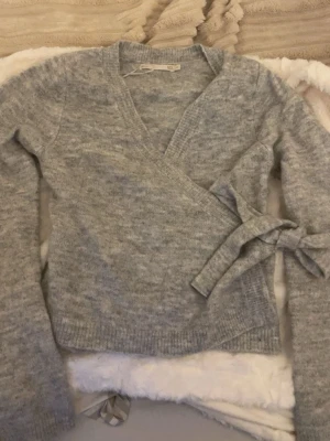 Grå omlottkofta från 157 XS - Mysig ljusgrå omlottkofta från 157 i storlek XS. Koftan har långa ärmar, ribbade muddar och knytband i sidan för en snygg passform. Perfekt till jeans eller kjol och superenkel att styla till olika looks.