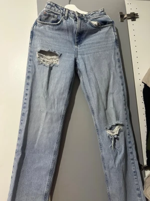 Ljusblå slitna straight jeans - Säljer ett par ljusblå jeans med raka ben och stora slitningar på både lår och knä. Klassisk femficksmodell i denim med hög midja och vintagekänsla. Perfekta för dig som gillar en avslappnad och trendig look.