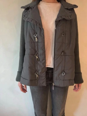 Mörkgrå quiltad dunväst med detaljer - Snygg mörkgrå quiltad dunväst med coola metallhakar framtill och en bred krage med volangkant. Västen har en rak passform och är tillverkad i ett mjukt, vadderat material som håller värmen. Perfekt att slänga över en stickad tröja för extra lager.