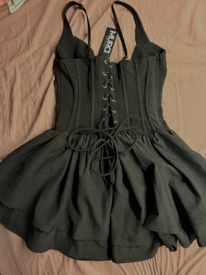 Ny Murci Layerd Black Mini Dress - Svart klänning med korsettliknande överdel och justerbara axelband. Figurnära upptill och lätt utsvängd kjol. Enkel och snygg modell som passar bra till fest eller utekväll.