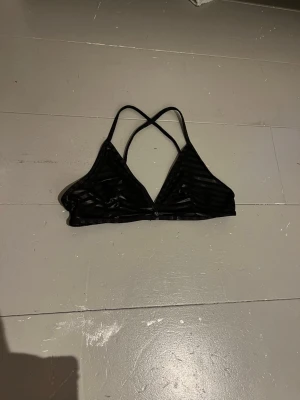 Svart randig bikini överdel med smala band - Säljer en svart bikini överdel med tunna axelband som korsas i ryggen. Bikini överdelen har ett randigt mönster i transparent mesh och en enkel, triangelformad design.