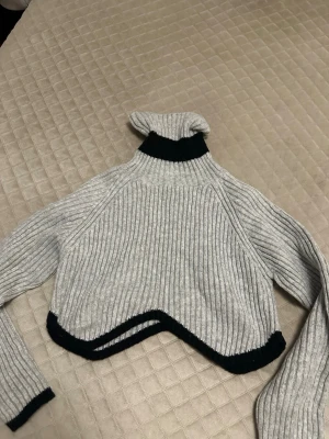 Grå croppad polotröja med svarta detaljer - Säljer en ribbstickad grå polotröja med svarta kanter på ärmslut, nederkant och krage. Tröjan är croppad med en unik vågig nederkant och har lång ärm och hög polokrage. Perfekt för dig som vill ha en trendig och mysig look.